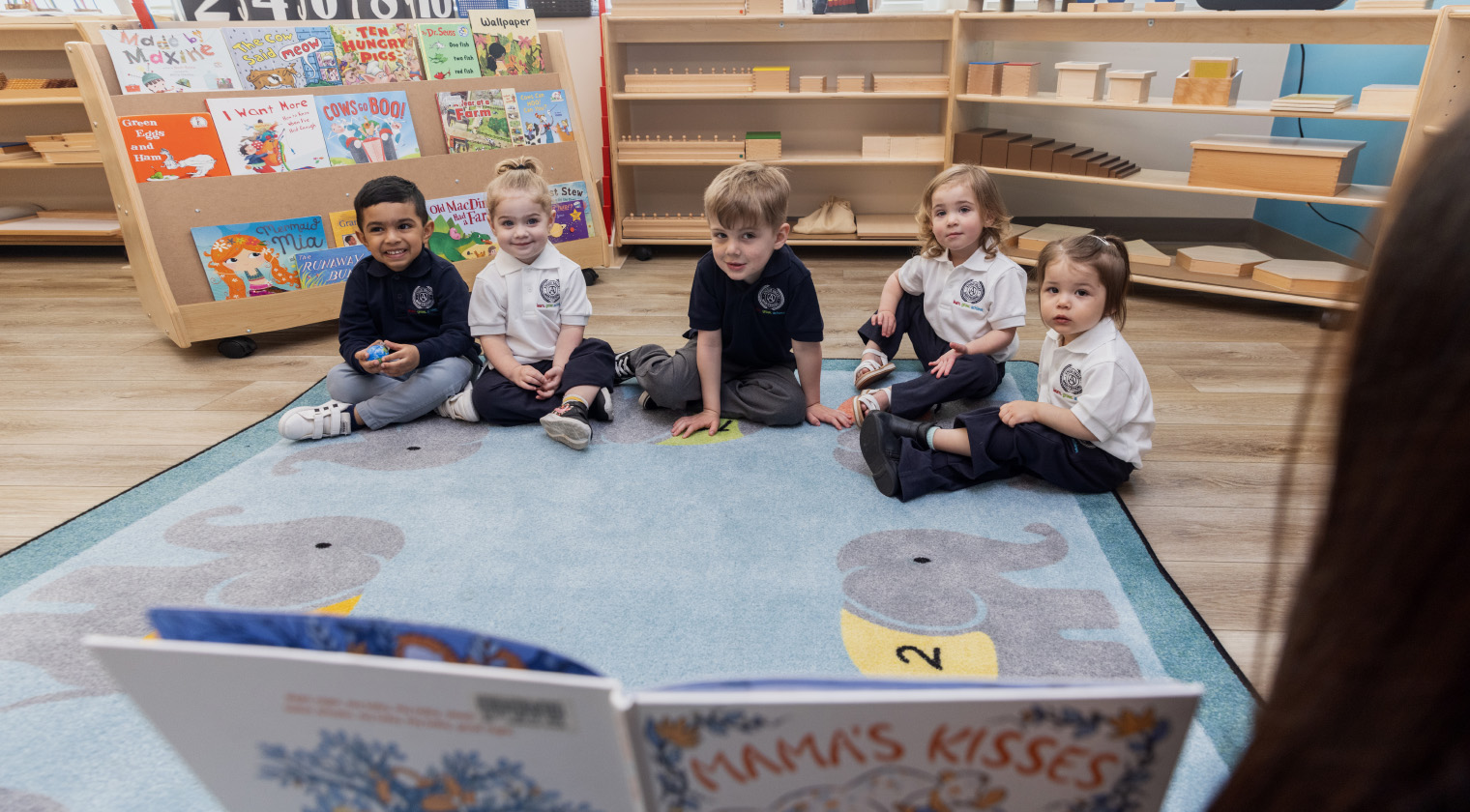 Five young children, of different ages and ethnicities, sit in a row on a light blue area carpet, shelves of books and Montessori materials are behind them, listening to a teacher reading aloud