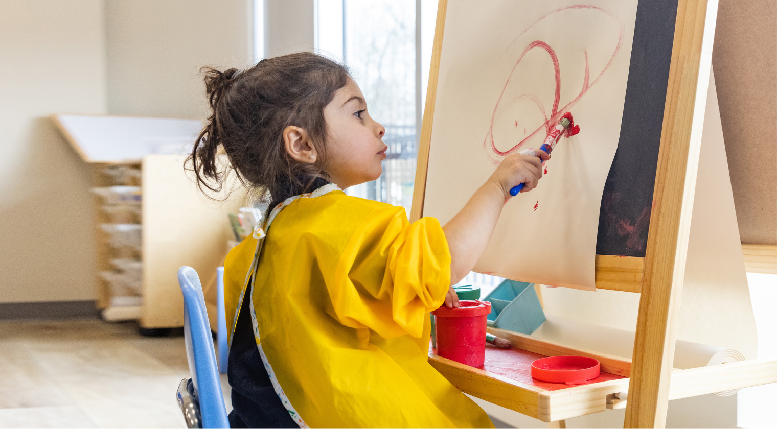 Little girl is seated by a classroom window at an art easel, wearing a bright yellow smock, painting in bright red